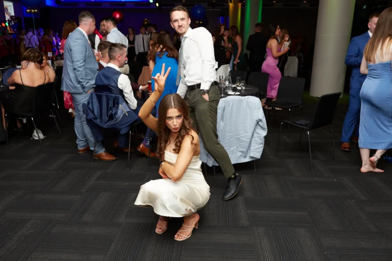 Candid moment at St. Kevin’s 65th Anniversary Ball at Marvel Stadium, showing a playful scene with a woman in a light-colored dress crouching and making a peace sign, while a man in a white shirt and dark pants stands behind her smiling. Other formally dressed guests mingle in the dimly lit background with patterned carpet and festive lighting. The image captures a relaxed, joyful, and celebratory atmosphere among attendees enjoying the milestone anniversary event.