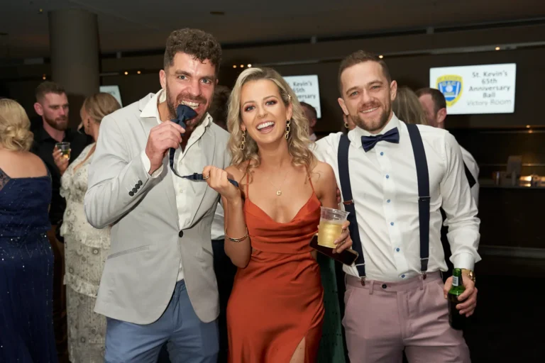 Candid moment at St. Kevin’s 65th Anniversary Ball at Marvel Stadium, showing three guests enjoying playful antics. A man on the left holds his bow tie in his mouth with a wide smile, a woman in a burnt-orange slip dress grins while holding a drink and another bow tie, and a man on the right in suspenders and light pink trousers raises his glass. The background features other guests and a partial banner, capturing the joyful, celebratory atmosphere and camaraderie of the milestone event.