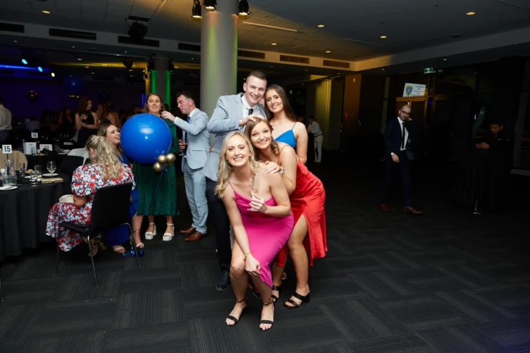 Candid moment at St. Kevin’s 65th Anniversary Ball at Marvel Stadium, featuring a group of young adults dressed in elegant formal attire—a grey suit, hot pink, vibrant red, and blue dresses—laughing and posing together. In the background, other guests mingle, with one playfully interacting with a large blue balloon. The image conveys joy, camaraderie, and the festive spirit of the milestone anniversary celebration.