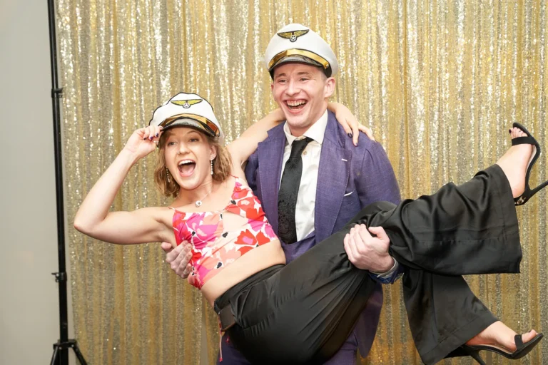 Candid moment at St. Kevin’s 65th Anniversary Ball in Marvel Stadium, showing two guests posing playfully in front of a gold sequin backdrop. A woman in a colorful top and dark pants, wearing a white pilot’s hat, is lifted in the arms of a man in a purple suit and matching pilot’s hat. Both laugh joyfully, creating a lively and memorable photo booth scene that highlights the fun, celebratory spirit of the evening.