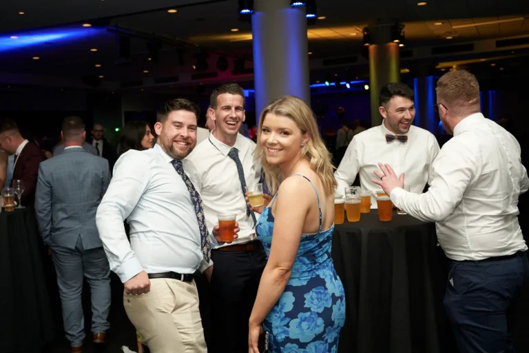 Candid moment at St. Kevin’s 65th Anniversary Ball at Marvel Stadium, showing four guests enjoying the celebration. Three men in shirts and ties and a woman in a blue floral dress stand around a high-top table, holding drinks and smiling at the camera. Other attendees are visible in the softly lit background, creating a lively, social atmosphere. The image conveys joy, camaraderie, and the festive spirit of the milestone anniversary event.
