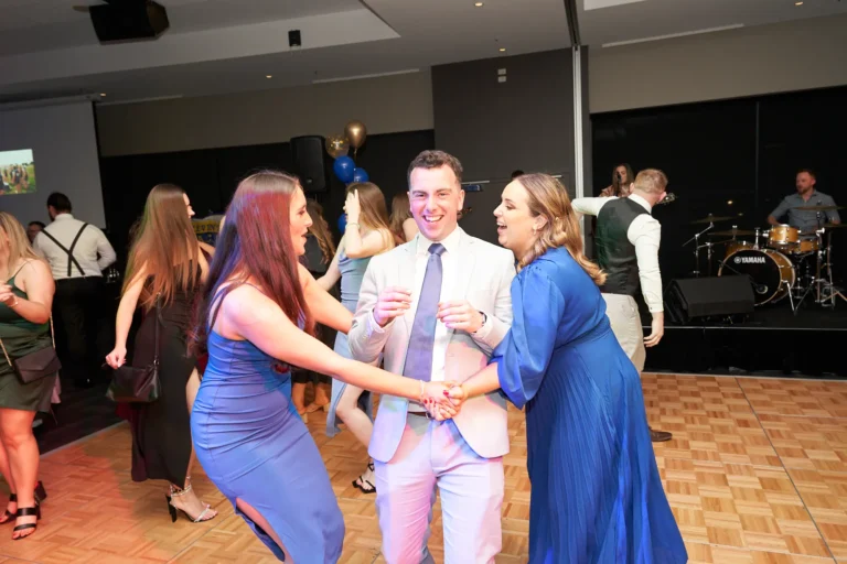 Guests dance joyfully on the lively floor of St. Kevin's 65th Anniversary Ball at Marvel Stadium, dressed in elegant formal attire. In the foreground, a man in a light-colored suit holds hands with two women in blue dresses, all smiling and immersed in the music. Behind them, other attendees enjoy the festivities while a live band, including a Yamaha drum set, provides entertainment. The dynamic lighting and energetic crowd create a vibrant, celebratory atmosphere, capturing the excitement, camaraderie, and unforgettable moments of this milestone anniversary event.