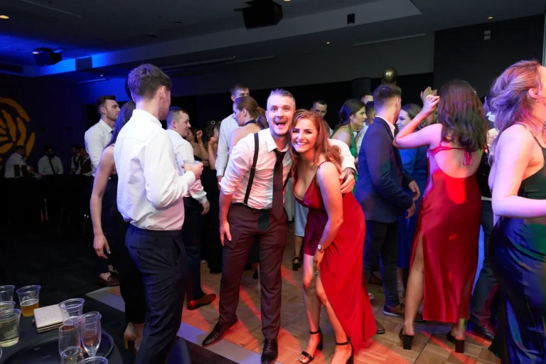 This image captures a lively moment on the dance floor at St. Kevin’s 65th Anniversary Ball at Marvel Stadium. A crowd of guests in formal attire is enjoying the festivities, with a prominent couple in the foreground smiling joyously. The man, wearing a white shirt and suspenders, embraces a woman in a striking red gown. Other attendees are seen mingling and dancing in the background, while dynamic lighting adds energy and a celebratory ambiance, conveying the joy and camaraderie of the milestone anniversary event.