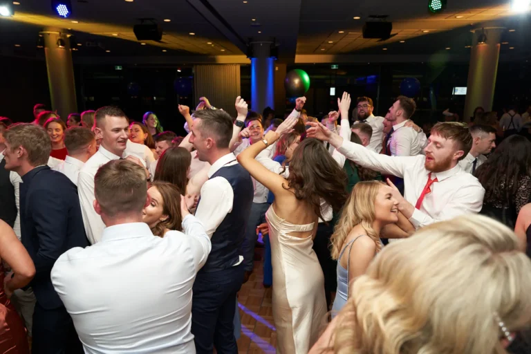 This image captures a lively moment on the dance floor at St. Kevin’s 65th Anniversary Ball at Marvel Stadium. A group of young adults in formal attire, including suits and elegant dresses, dance enthusiastically with smiles and raised hands. The dimly lit venue, festive decorations, and dynamic interactions among guests create an energetic, joyful atmosphere, reflecting the camaraderie and celebratory spirit of this milestone anniversary event.