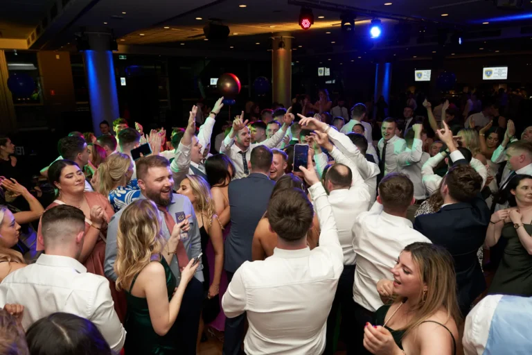This image captures a lively moment on the dance floor at St. Kevin’s 65th Anniversary Ball at Marvel Stadium. A large group of elegantly dressed attendees are dancing energetically, raising their hands, and some capturing the moment on their phones. Blue and purple atmospheric lighting fills the formal event space, enhancing the festive ambiance. Guests are smiling, laughing, and interacting with each other, reflecting the joy, camaraderie, and high-energy celebration of this milestone anniversary event.
