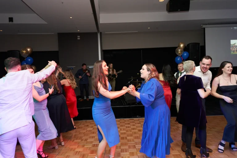 Guests celebrate on the dance floor at St. Kevin's 65th Anniversary Ball at Marvel Stadium, dressed in elegant suits and dresses. In the foreground, individuals hold hands in a lively group dance, smiling and enjoying the moment. Blue and gold balloons add festive charm, while a live band performs on stage in the background. The image captures the joyous energy, camaraderie, and vibrant atmosphere of the milestone anniversary celebration.
