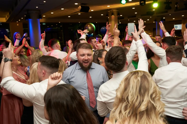 This image captures a vibrant moment on the dance floor at St. Kevin’s 65th Anniversary Ball at Marvel Stadium. A bearded man in a checked shirt and red tie smiles broadly with his hands raised, surrounded by jubilant guests dancing and celebrating. Many attendees also raise their hands or capture the moment on phones, while colorful lighting illuminates the lively event space. The crowd’s expressions and interactions convey high energy, joy, and camaraderie, reflecting the festive and dynamic atmosphere of this milestone anniversary celebration.
