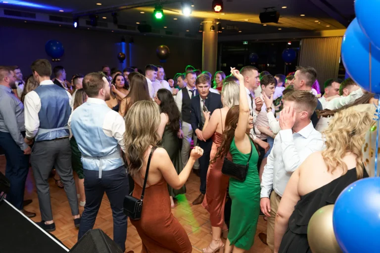 This image captures a lively moment on the dance floor at St. Kevin’s 65th Anniversary Ball at Marvel Stadium. Guests in elegant dresses and suits are dancing, laughing, and socializing under colorful party lights. Balloons and dynamic lighting create a festive atmosphere, while attendees raise their hands and engage with the music, reflecting the joyous energy and celebratory spirit of this milestone anniversary event.
