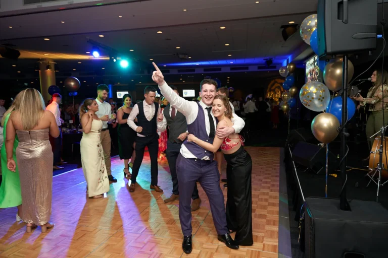 Guests enjoy a lively moment on the dance floor at St. Kevin’s 65th Anniversary Ball. In the foreground, a man in a white shirt and tie points upward while a woman in a black and red outfit embraces him, both smiling. Other attendees dance and socialize on the illuminated floor, with blue and gold balloons decorating the background. A band set-up with instruments is visible, enhancing the festive and celebratory atmosphere of the anniversary ball.