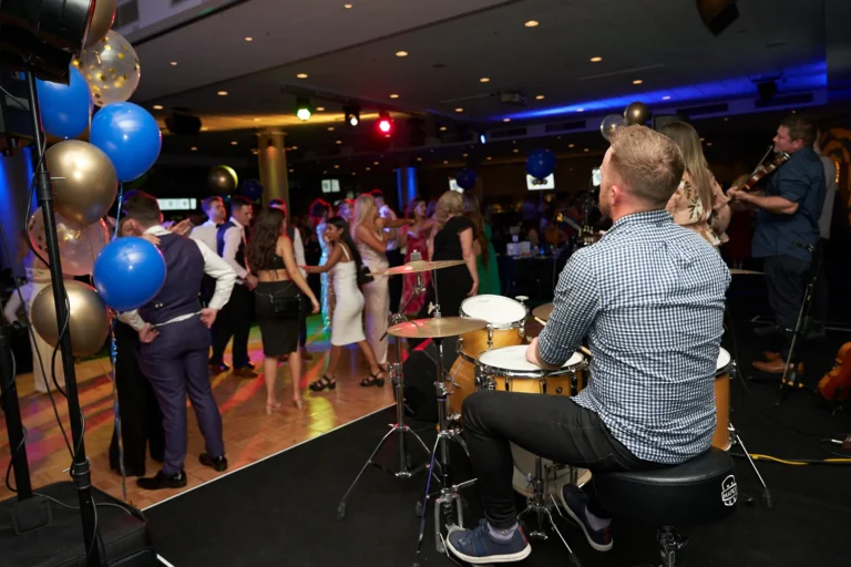 A lively dance floor scene at St. Kevin's 65th Anniversary Ball features a drummer in a plaid shirt playing a drum kit, while a band member behind plays a string instrument. Elegantly dressed guests, including men in suits and women in formal attire, enjoy dancing and mingling under festive lights. Blue and gold balloons add a celebratory touch, capturing the joyous energy and live music atmosphere of the anniversary ball.