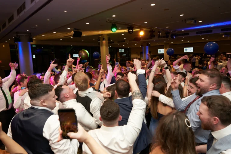 This image captures a vibrant moment on the dance floor at St. Kevin’s 65th Anniversary Ball at Marvel Stadium. Guests, mostly men in formal attire with some women, dance energetically with hands raised, expressing joy and camaraderie. Colorful lighting, including green and red spotlights, illuminates the room alongside festive balloons, creating a lively and celebratory atmosphere. The dynamic crowd, smiling and fully engaged in the music, reflects the electric energy and social spirit of this milestone anniversary event.