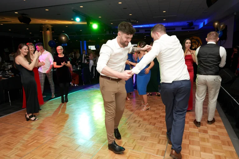 Two men, dressed in white shirts and formal trousers, dance joyfully on the polished wooden floor at St. Kevin's 65th Anniversary Ball at Marvel Stadium, holding hands and smiling broadly. Around them, guests in elegant gowns and suits watch, clap, and interact, adding to the lively energy. Blue and gold balloons float above the dance floor, while ambient lighting illuminates the spacious event hall. In the background, musicians and a stage setup hint at live entertainment. The scene captures the festive spirit, camaraderie, and jubilant atmosphere of this milestone anniversary celebration.