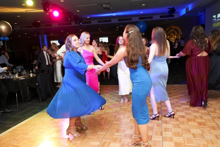 A lively scene on the dance floor at St. Kevin's 65th Anniversary Ball shows elegantly dressed women holding hands and dancing, their colorful gowns swirling with movement. The foreground features a woman in a vibrant blue dress smiling at the camera, while another in a pink dress dances beside her. Guests mingle at tables in the background, with festive balloons and colorful lighting adding to the celebratory ambiance. The polished wooden floor reflects the lights and energy, capturing the joyful and dynamic atmosphere of the anniversary celebration.