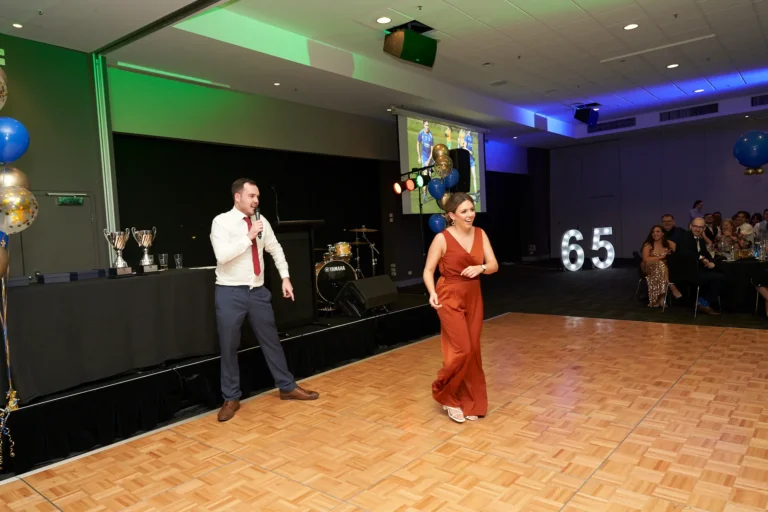 This photo from St. Kevin’s 65th Anniversary Ball at Marvel Stadium captures a lively moment on the polished wooden dance floor. A man in a white shirt holds a microphone, likely serving as the emcee, while a woman in a reddish-brown jumpsuit moves nearby, adding energy to the scene. In the background, a stage with drums, trophies, and a large projection screen sets the atmosphere, highlighted by an illuminated “65” sign and festive balloons. Guests seated at decorated tables complete the celebratory setting, blending formal recognition with entertainment and joyous camaraderie.