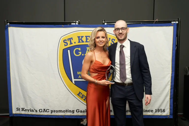This image captures a proud moment at St. Kevin’s 65th Anniversary Ball at Marvel Stadium, showing a woman in a striking orange gown receiving an award from a male presenter in a dark suit. She holds the trophy with a beaming smile, while a large banner behind them reads “St. Kevin’s G.A.C. promoting Gael since 1956,” emphasizing the club’s long-standing legacy. The image conveys a sense of achievement, celebration, and the formal, festive atmosphere of the milestone anniversary event.