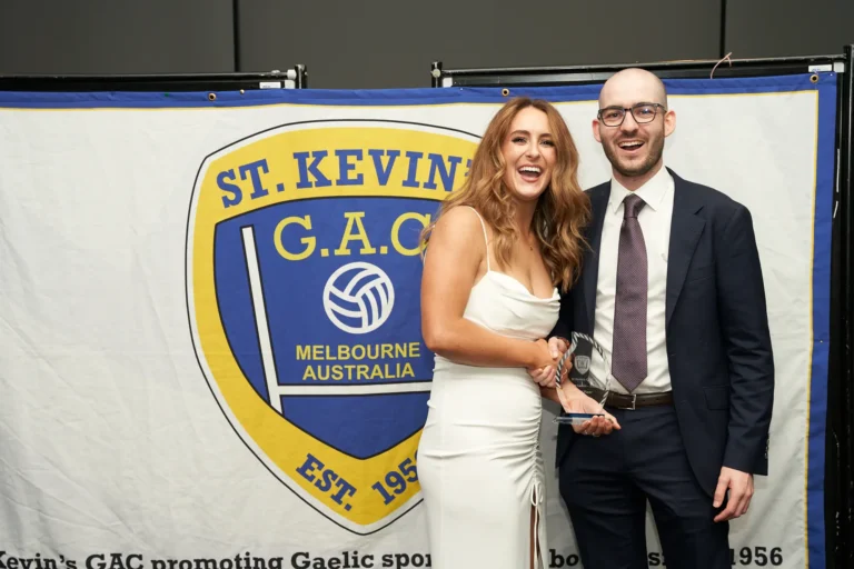 This image captures a joyful moment at St. Kevin’s 65th Anniversary Ball at Marvel Stadium, where a smiling woman in a white dress proudly holds her award. Beside her, a man in a suit, likely the presenter, stands beaming in celebration. Behind them, a banner prominently displays the St. Kevin’s G.A.C. crest with “Melbourne Australia Est. 1956,” highlighting both the significance of the award presentation and the club’s enduring history. The scene reflects pride, recognition, and the festive spirit of the milestone event.