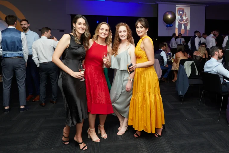 This image captures four elegantly dressed women at St. Kevin’s 65th Anniversary Ball at Marvel Stadium, smiling and posing for the camera while holding drinks. They wear formal gowns in black, red, grey, and golden yellow. In the background, guests in formal attire mingle across a spacious ballroom, with a large screen displaying a crest or emblem, emphasizing the celebratory setting. The scene conveys a lively, elegant, and convivial atmosphere, highlighting the joy and camaraderie of attendees at this milestone anniversary gala.