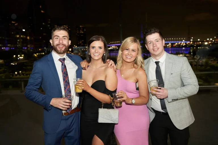 This image captures a joyful moment at St. Kevin’s 65th Anniversary Ball at Marvel Stadium, featuring two couples dressed in elegant evening attire. The men wear a blue suit with patterned tie and a grey suit with black tie, while the women are in a black strapless dress and a pink one-shoulder gown. All four are smiling and holding drinks, enjoying the celebratory atmosphere. The dimly lit background with distant city lights suggests an evening outdoor or semi-outdoor setting, highlighting the festive and lively ambiance of the milestone anniversary event.
