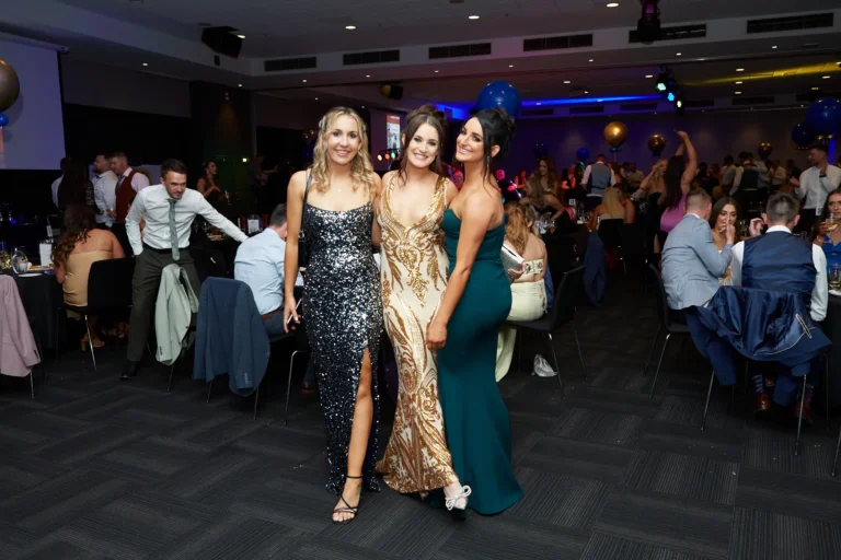 This image captures a glamorous moment at St. Kevin’s 65th Anniversary Ball at Marvel Stadium, featuring three women in elegant full-length gowns—a dark sequined dress with a thigh-high slit, a gold sequined gown, and an emerald green strapless dress—smiling and posing for the camera. The background shows a large banquet hall with tables set for dining, blue balloons, and formally dressed guests mingling, highlighting the festive and celebratory atmosphere of the milestone anniversary event. The scene exudes joy, sophistication, and camaraderie among attendees.