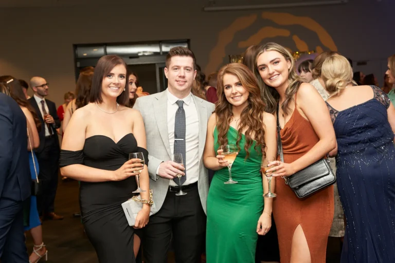 This image captures a cheerful group moment at St. Kevin’s 65th Anniversary Ball at Marvel Stadium. Four elegantly dressed individuals pose together, with two women on the left in a black strapless gown and a vibrant green dress, a man in a grey suit with patterned tie in the center, and another woman in a rust-colored gown on the right. Several hold celebratory drinks, and all are smiling toward the camera. The festive background, with other guests and warm lighting, conveys the joyful and lively atmosphere of this milestone celebration.