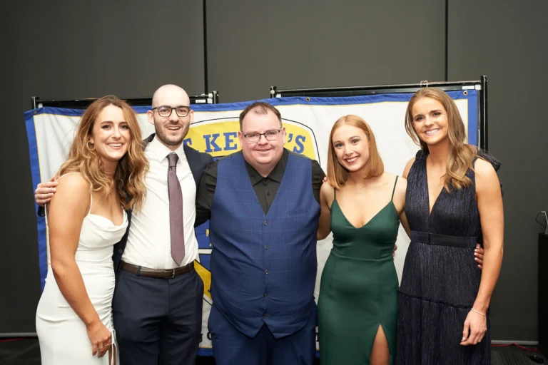 This image captures a cheerful group moment at St. Kevin’s 65th Anniversary Ball at Marvel Stadium. Five elegantly dressed guests pose together, including three women in formal gowns—white, green, and dark blue—and two men in suits, one wearing a blue vest. They smile warmly at the camera, exuding a festive and celebratory atmosphere. A partially visible banner in the background likely referencing “St. Kevin’s” adds context to the milestone event, highlighting camaraderie and joy among friends and colleagues enjoying the anniversary celebration.