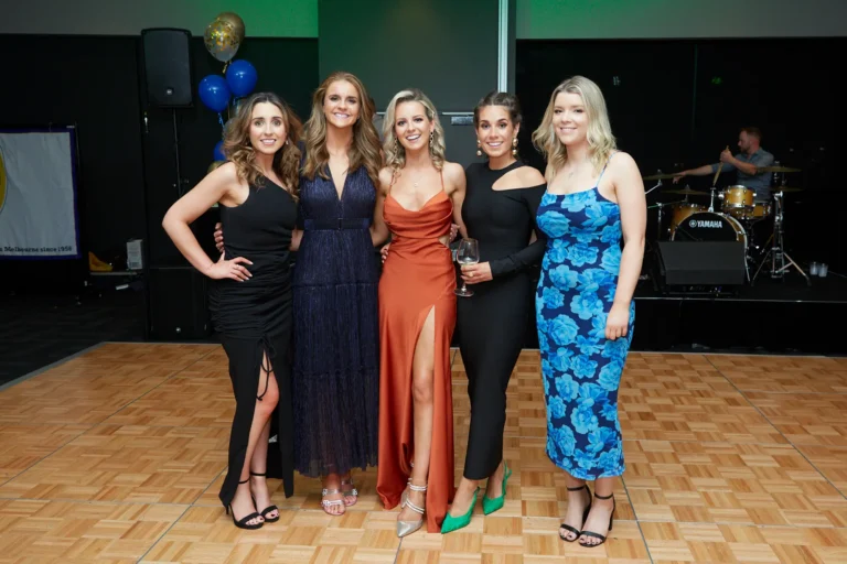 This image captures a lively group moment at St. Kevin’s 65th Anniversary Ball at Marvel Stadium. Five elegantly dressed women pose together on a dance floor, smiling and enjoying the celebratory atmosphere. Their formal gowns vary in color and style, adding vibrancy to the scene. In the background, a drummer is visible behind a Yamaha drum kit, suggesting live music and entertainment at the event. The overall composition conveys joy, camaraderie, and the festive spirit of the milestone anniversary celebration.