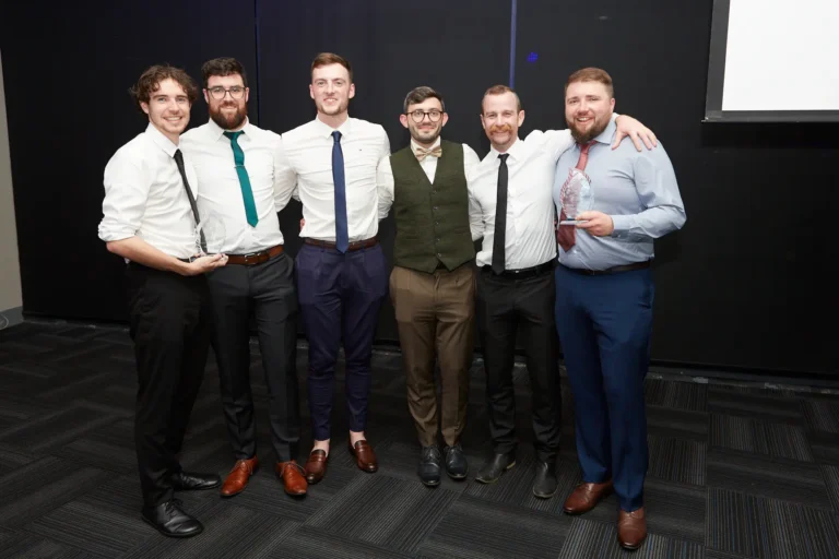 This image captures a celebratory moment at St. Kevin’s 65th Anniversary Ball at Marvel Stadium, featuring six formally dressed men posing with pride. Two of the men prominently display their awards, highlighting recognition and achievement during the milestone event. Attire includes a mix of white shirts, ties, bow ties, vests, and suits, reflecting the formal nature of the gala. The dark backdrop and patterned flooring provide a professional setting, while the expressions and posture of the group convey camaraderie, accomplishment, and the festive spirit of the anniversary celebration.