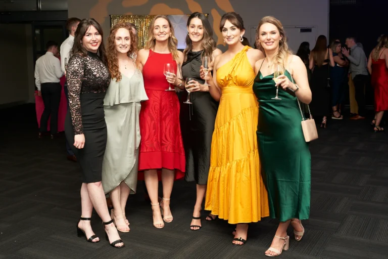 This image captures a lively group moment at St. Kevin’s 65th Anniversary Ball at Marvel Stadium. Six elegantly dressed women pose together, holding drinks and smiling broadly, exuding joy and camaraderie. Their formal dresses feature a range of colors including black, red, yellow, green, and light grey, reflecting the celebratory nature of the occasion. The softly blurred background suggests a bustling gala atmosphere, highlighting the social and festive spirit of the milestone anniversary event.