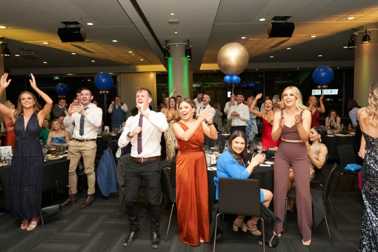This wide-angle photo from St. Kevin’s 65th Anniversary Ball at Marvel Stadium captures candid guest reactions to an award winner. Attendees in formal attire are seated at round tables, clapping, cheering, and raising their hands in excitement, their expressions filled with joy and surprise. Prominent gold and blue balloons enhance the festive atmosphere, highlighting the celebratory spirit of the milestone evening.