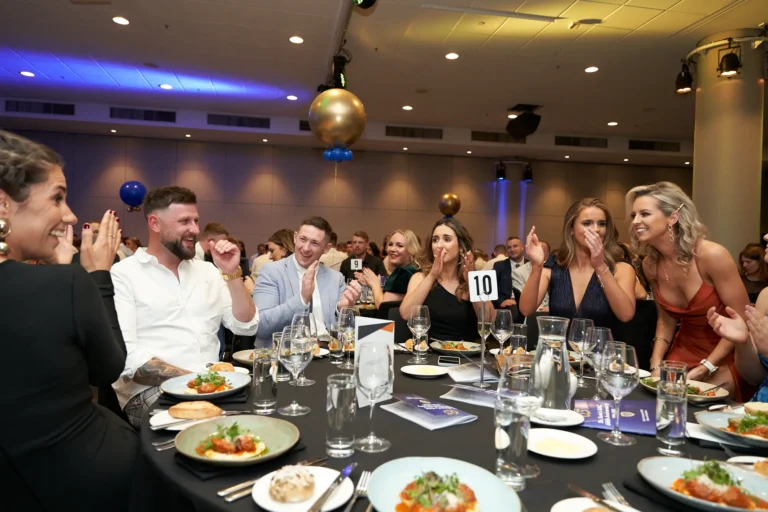 This image from St. Kevin’s 65th Anniversary Ball at Marvel Stadium captures a celebratory moment during the awards presentation. At an elegantly set guest table, a woman begins to rise from her seat, preparing to walk toward the stage as fellow attendees in formal attire smile, applaud, and react with excitement. The table is adorned with glasses, plates, and details of a multi-course meal, reflecting the gala dinner setting. In the background, other guests and a large golden balloon add to the festive atmosphere, highlighting the joy and anticipation of this milestone celebration.