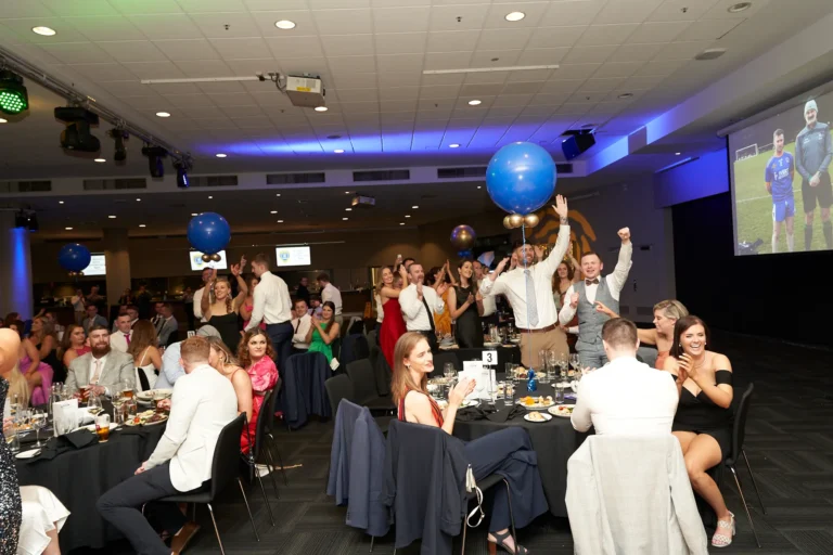 This wide-angle photo from St. Kevin’s 65th Anniversary Ball at Marvel Stadium captures candid guest reactions during the awards ceremony. Guests, elegantly dressed in formal attire, are seated around black-draped tables, while some cheer and raise their arms in celebration. A large screen in the background displays award-related visuals, and festive blue balloons add to the jubilant atmosphere. The scene conveys the excitement, camaraderie, and celebratory spirit of the milestone event.