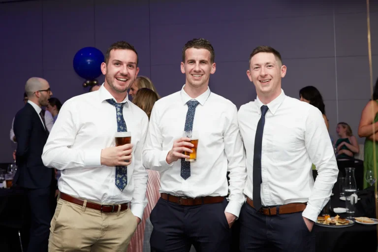 This image captures three men at St. Kevin’s 65th Anniversary Ball at Marvel Stadium, dressed in white shirts and patterned blue ties, smiling and holding glasses of beer. They stand together in a formal event setting, with other guests and tables visible in the softly lit background. A large blue balloon adds a festive touch, enhancing the celebratory atmosphere. The candid moment conveys camaraderie and enjoyment among attendees during the milestone anniversary celebration.