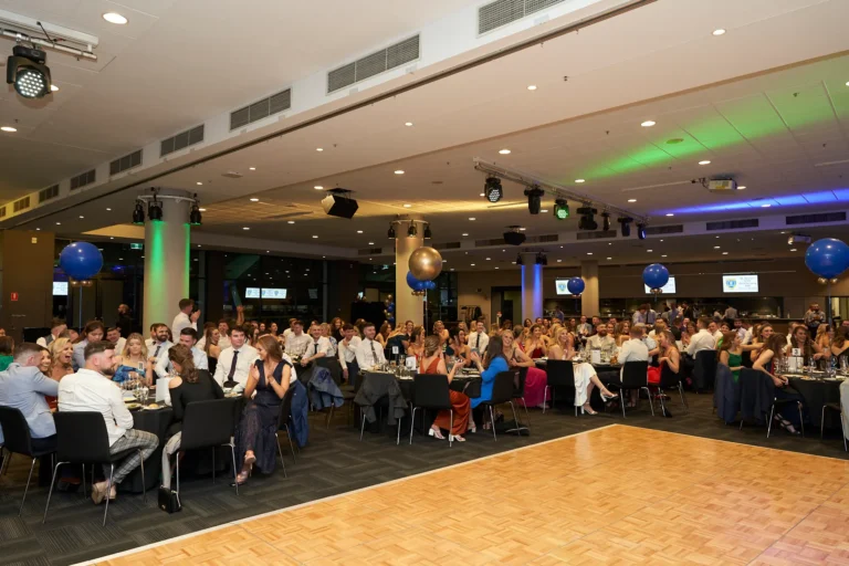 Guests enjoy the presentation segment at St. Kevin’s 65th Anniversary Ball at Marvel Stadium, seated at round tables in a ballroom decorated with large blue and gold balloons. Stage lighting casts vibrant green and blue hues across the room, while attendees in formal attire engage in conversation and observe the presentation, capturing the lively and celebratory atmosphere of this milestone anniversary event.