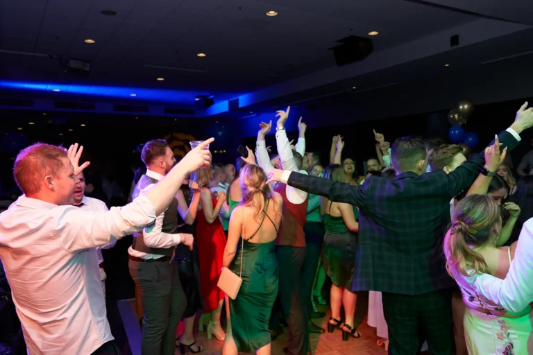 Guests celebrate on the dance floor at St. Kevin’s 65th Anniversary Ball, dressed in formal attire. Attendees laugh, dance, and interact under vibrant lighting, with blue and gold balloons enhancing the festive and joyful atmosphere of the milestone celebration.