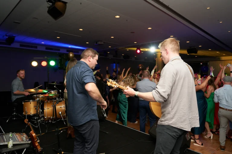 This image captures a vibrant moment on the dance floor at St. Kevin’s 65th Anniversary Ball at Marvel Stadium. A live band, including a drummer and guitarist, performs on stage in the foreground, while guests fill the dance floor in the background, dancing with raised hands and joyful expressions. Colorful, festive lighting and balloons enhance the celebratory atmosphere, creating a lively and energetic scene. The photograph highlights the excitement, music, and camaraderie of the anniversary ball, perfectly showcasing the memorable moments and joyous interactions of attendees during the milestone celebration.