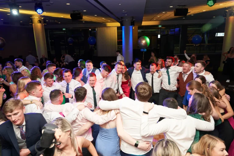 This overhead wide shot captures a lively moment on the dance floor at St. Kevin’s 65th Anniversary Ball at Marvel Stadium. A large group of guests, dressed in formal attire including suits, ties, and elegant dresses, dance and socialize with joy. Arms are linked and smiles beam across faces, reflecting camaraderie and celebration. Festive lighting illuminates the room, enhancing the energetic and dynamic atmosphere. The image conveys the collective happiness and spirited energy of the milestone anniversary, showcasing both the vibrant dance floor and the celebratory ambiance of the event.