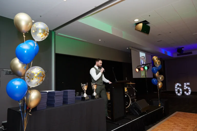 This image from St. Kevin’s 65th Anniversary Ball at Marvel Stadium captures a male presenter in a vest and shirt addressing the audience from a podium. To the side of the stage, a decorative display includes bunches of blue and gold balloons, gleaming trophies, and stacks of certificates awaiting presentation. Behind the presenter, musical instruments and a lit “65” add to the celebratory setting, blending awards and entertainment in one memorable evening.
