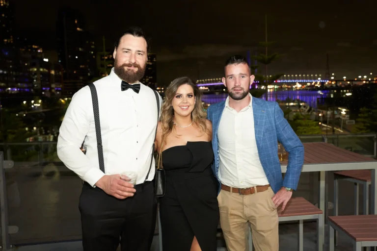 This image captures three guests at St. Kevin’s 65th Anniversary Ball at Marvel Stadium, enjoying the evening on an outdoor terrace. A bearded man on the left wears a white shirt with suspenders and a black bow tie, holding a drink. The central woman wears a strapless black gown with a side slit and a gold chain bag, smiling at the camera. To the right, a man in a blue blazer and beige trousers stands casually with one hand in his pocket. City lights and a waterfront in the background create an elegant, celebratory atmosphere.