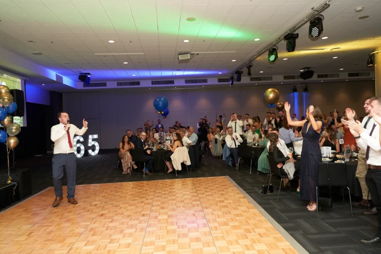 This wide-angle image from St. Kevin’s 65th Anniversary Ball at Marvel Stadium captures a lively moment as a speaker addresses the audience from the wooden dance floor. His hand is raised emphatically, emphasizing his speech, while guests seated at numerous round tables respond with applause and engaged expressions. Blue and gold balloons decorate the spacious room, adding a festive touch. The photograph effectively conveys the energy, attentiveness, and celebratory atmosphere of the milestone anniversary event, highlighting the connection between the speaker and the audience.