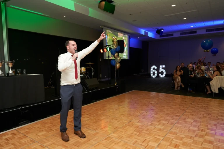This image from St. Kevin’s 65th Anniversary Ball at Marvel Stadium captures a dynamic moment on stage, where a speaker in a white shirt and tie passionately addresses the audience with a microphone. Behind him, the large illuminated “65” highlights the milestone celebration. The polished wooden stage contrasts with the carpeted floor filled with elegantly set round tables, decorated with balloons and formal place settings. Guests seated at the tables appear engaged and attentive, contributing to the festive and commemorative atmosphere of this special anniversary event.