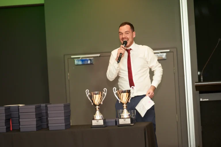 A male speaker in a white shirt and red tie addresses the audience from a podium during the Presentation & Awards segment of St. Kevin’s 65th Anniversary Ball at Marvel Stadium. Two gleaming trophies and stacks of awards rest on a draped table beside him, hinting at upcoming recognitions. The background features modern doors and subtle lighting, while the foreground shows guest tables, capturing the formal, celebratory, and engaging atmosphere of the milestone anniversary event.