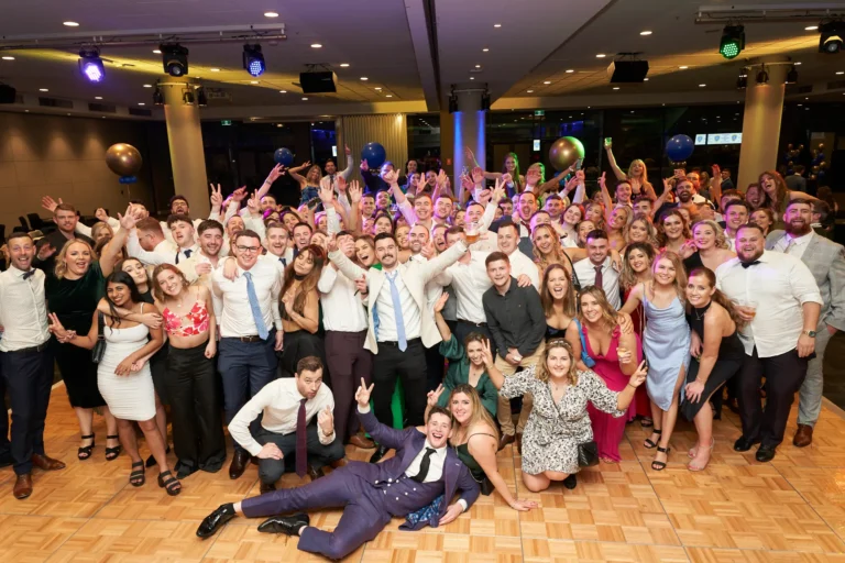 This wide-angle image captures a lively group of attendees at St. Kevin’s 65th Anniversary Ball, posing on a polished dance floor. Guests, dressed in elegant gowns and suits, smile, laugh, and raise their hands in celebration. Decorative balloons and ambient evening lighting enhance the festive atmosphere, while the large group conveys the joyous, communal spirit of the milestone anniversary event.