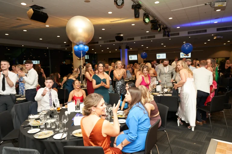 A wide-angle view of guests enjoying the awards presentation at St. Kevin’s 65th Anniversary Ball at Marvel Stadium. Attendees in formal attire are seated at elegantly decorated tables, interacting, cheering, and celebrating their friends’ achievements. Large gold and blue balloons float above the room, adding a festive touch. The image captures the lively energy, camaraderie, and joyful spirit of this milestone anniversary event.