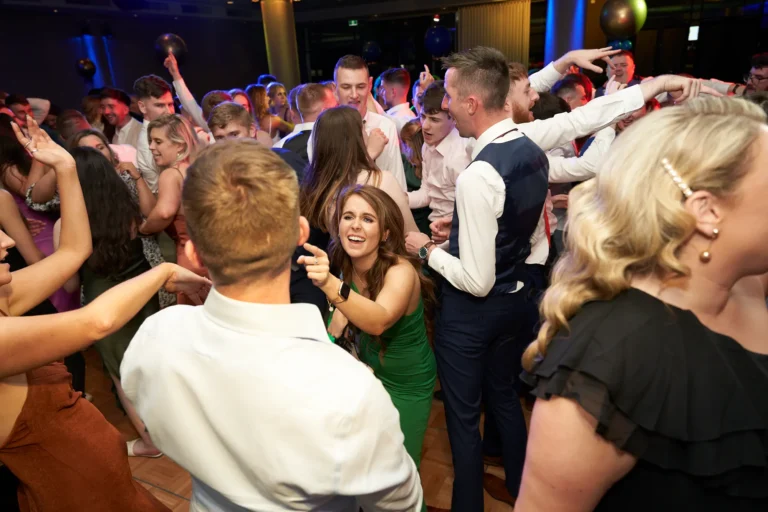 This wide shot captures a lively and energetic moment on the dance floor at St. Kevin’s 65th Anniversary Ball at Marvel Stadium. A diverse group of guests in formal and semi-formal attire, including suits and elegant dresses, are dancing, laughing, and interacting with one another. The festive lighting and joyful expressions convey a spirited atmosphere of camaraderie and celebration, perfectly reflecting the energy and significance of this milestone anniversary event.