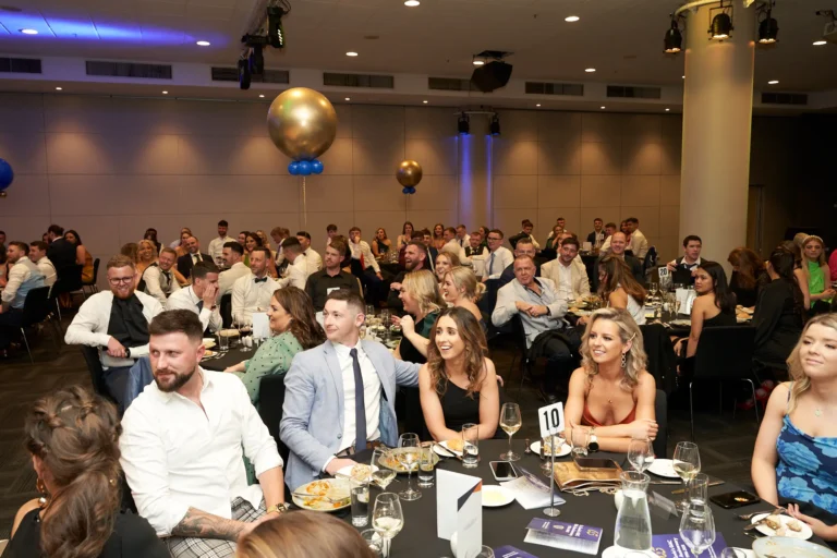 Guests enjoy candid moments at elegantly set tables during a presentation at St. Kevin’s 65th Anniversary Ball at Marvel Stadium. Attendees in formal attire engage in conversation and laughter, while golden balloons add a festive touch to the spacious hall. A large screen or stage in the distance highlights the focus of the event, capturing the celebratory atmosphere, communal spirit, and milestone significance of this anniversary gathering.