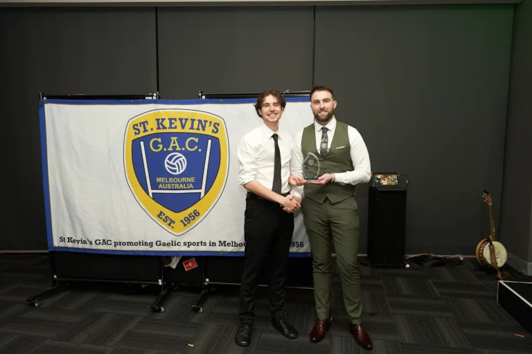 Award recipient shaking hands with the presenter at St. Kevin’s 65th Anniversary Ball, Marvel Stadium, Melbourne. The scene features a celebratory banner in the background highlighting the club’s history since 1956 and its role in promoting Gaelic sports, capturing the festive and formal atmosphere of the milestone event.