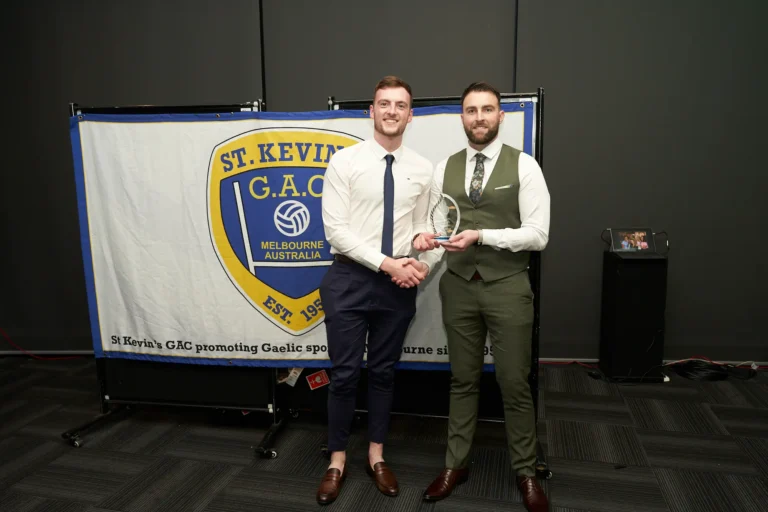 ward winner shaking hands with the presenter at St. Kevin's 65th Anniversary Ball at Marvel Stadium, with the club's banner and celebratory atmosphere highlighting the milestone event.
