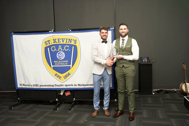 A presenter in a dark suit and waistcoat hands a clear trophy to a smiling recipient in a light-colored jacket and blue trousers at St. Kevin’s 65th Anniversary Ball at Marvel Stadium. Both stand in front of a large banner displaying the “ST. KEVIN'S G.A.C. MELBOURNE AUSTRALIA EST. 1956” logo, celebrating Gaelic sports in Melbourne. The formal attire, award, and banner emphasize the prestige and joyous atmosphere of this milestone anniversary celebration.
