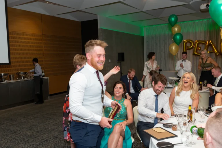 A joyful moment at the Pearses 50th Anniversary Awards Night at Zinc, Fed Square. A bearded man, wearing a white shirt and light vest, walks past a table holding a framed award, smiling broadly. Guests at the table, including a woman in green and two in white shirts, laugh and celebrate his achievement with raised hands. The background shows round tables with white tablecloths, glasses, and green and gold balloons spelling "PEA." The lively, festive atmosphere highlights the crowd’s warmth and the winner’s shared sense of accomplishment.