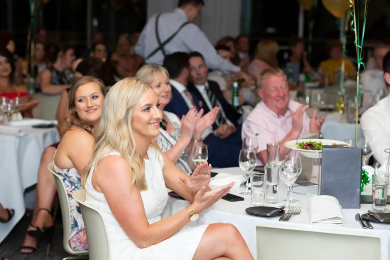 A lively moment at the Pearses 50th Anniversary Awards Night at Zinc, Fed Square, shows guests reacting with applause. In the foreground, a blonde woman in a white dress and another woman in a floral dress are smiling and clapping. The background features a formal dining hall with round tables, white tablecloths, green and gold balloons, and streamers. Guests at other tables are also clapping, while a waiter attends to guests in the distance. The scene captures the joyful and celebratory atmosphere of the awards presentation.